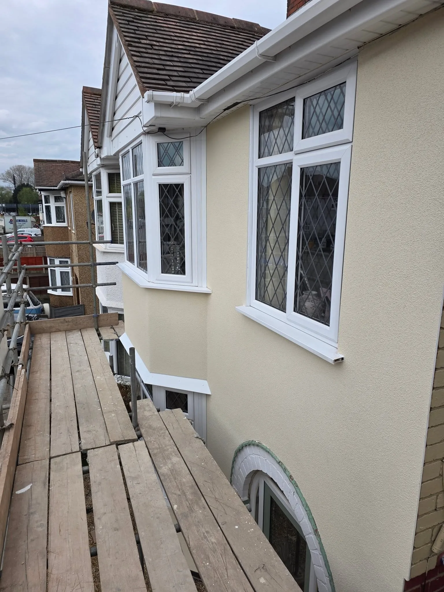 A finished house elevation in smooth cream render with a bay window, seen from the scaffold alongside.