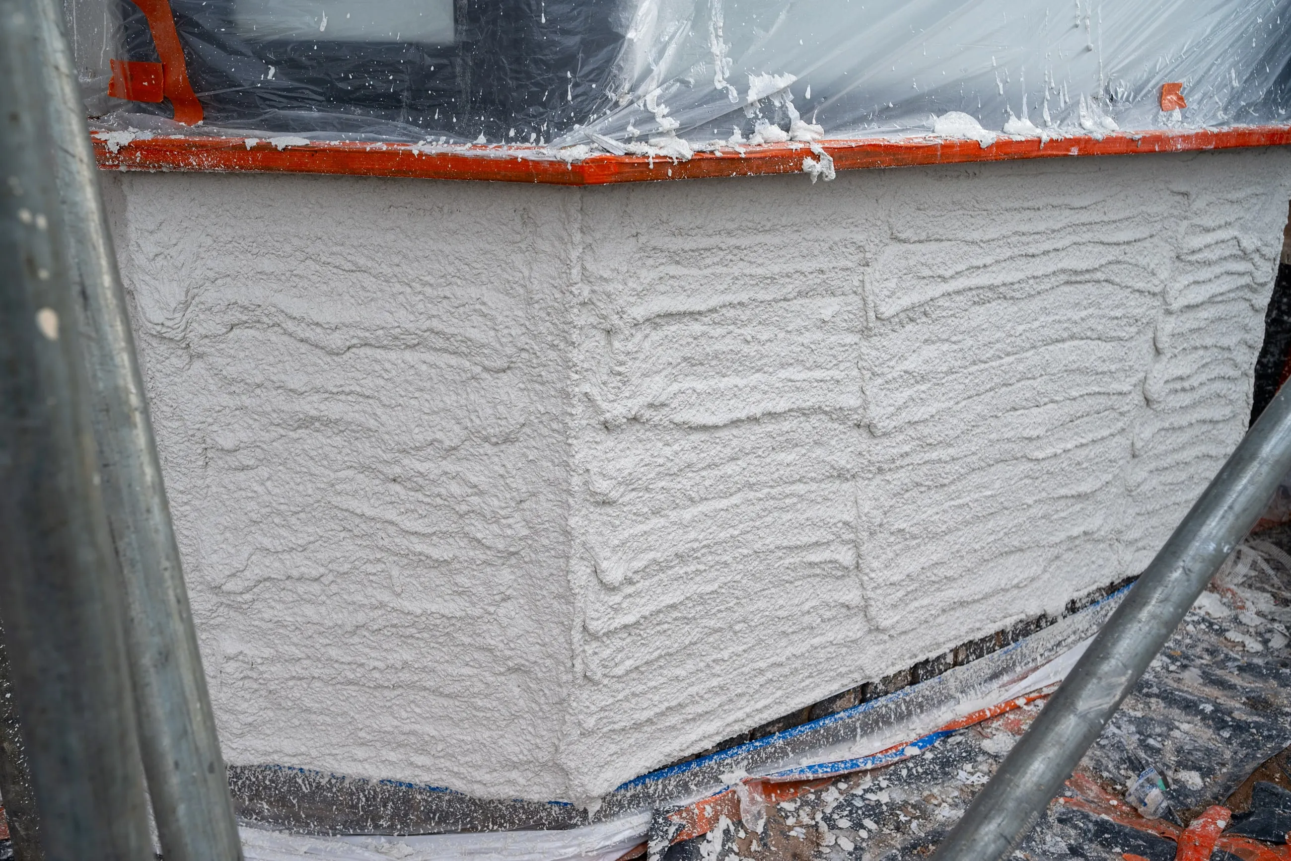 A close view of freshly applied white textured monocouche render on a wall corner, scaffolding visible alongside.